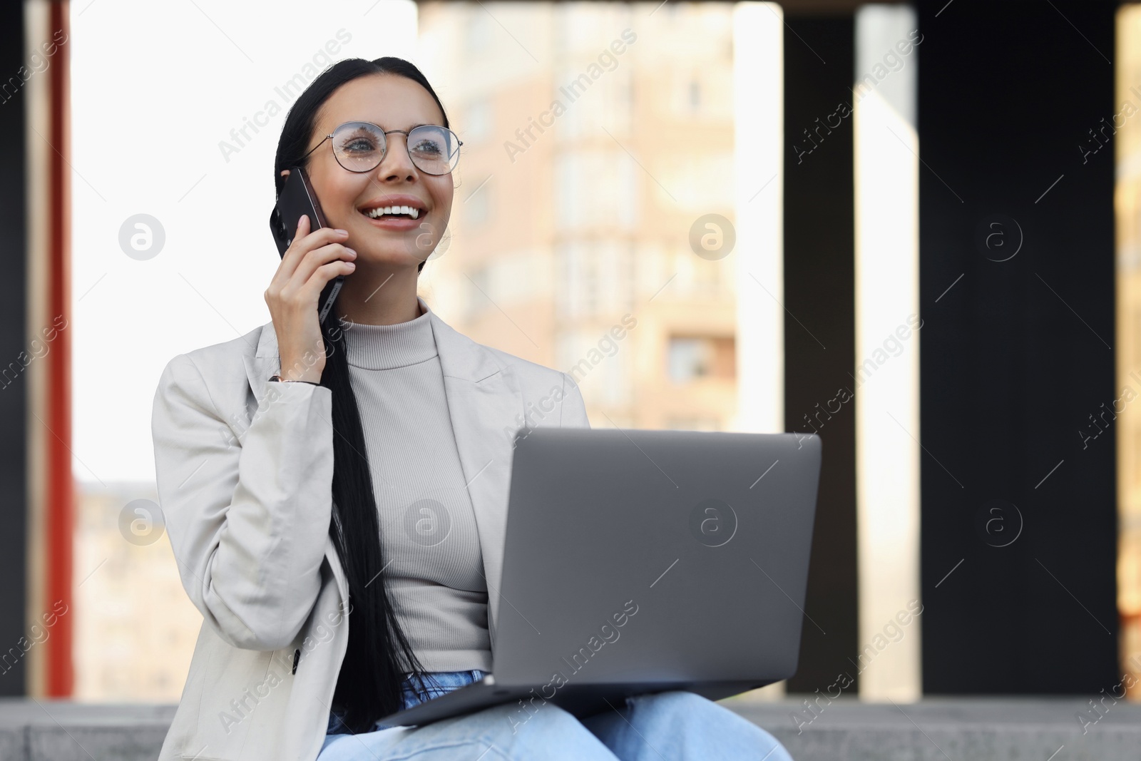 Photo of Happy young woman using modern laptop and talking on smartphone outdoors