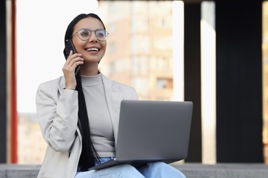 Photo of Happy young woman using modern laptop and talking on smartphone outdoors