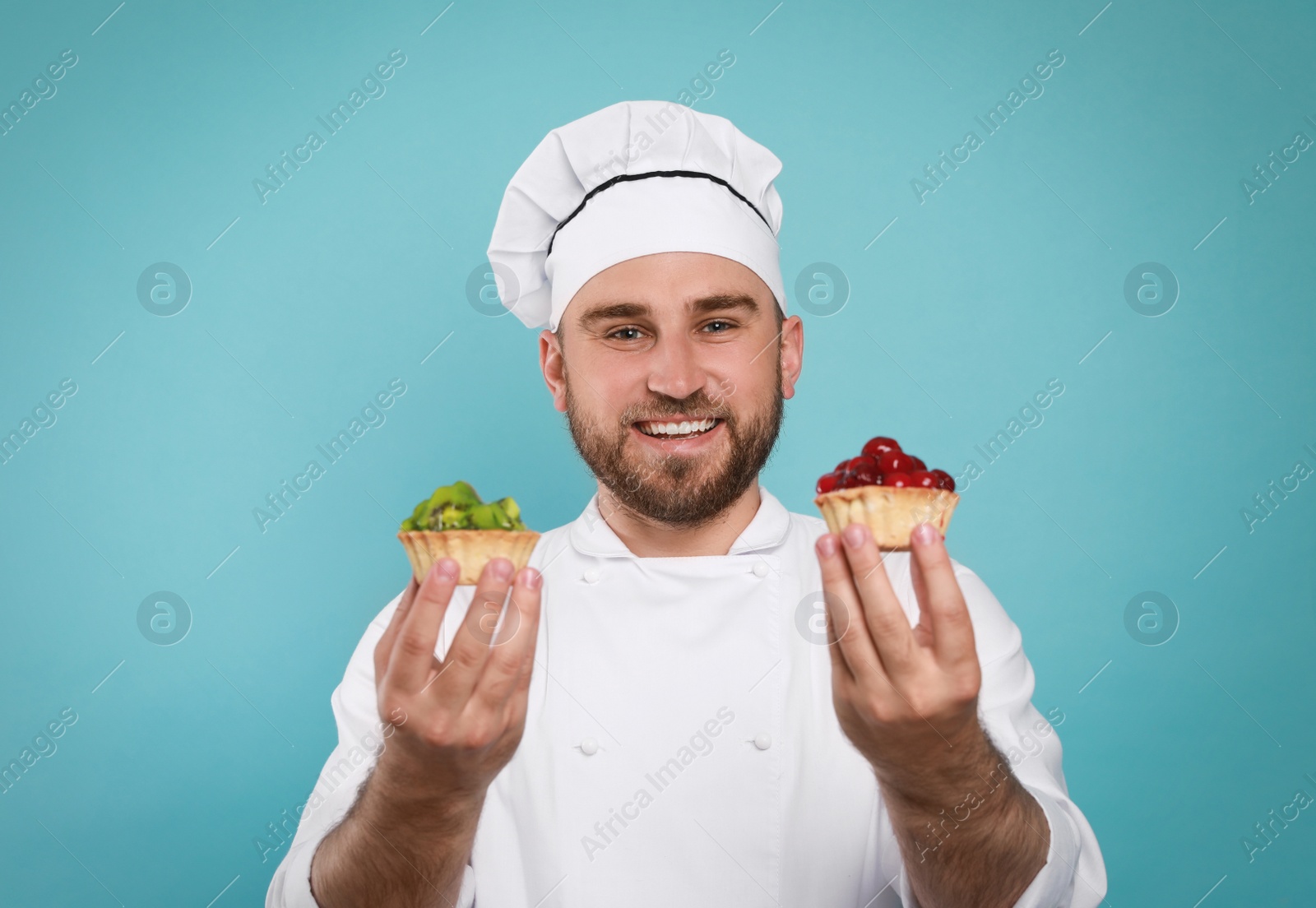 Photo of Happy professional confectioner in uniform holding delicious tartlets on light blue background