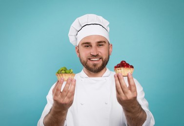 Happy professional confectioner in uniform holding delicious tartlets on light blue background