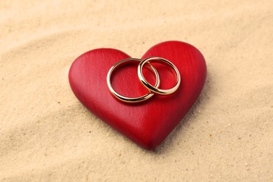 Honeymoon concept. Two golden rings and red wooden heart on sand