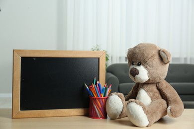 Teddy bear, small blackboard, pencils and pens in holder on wooden table indoors. Space for text