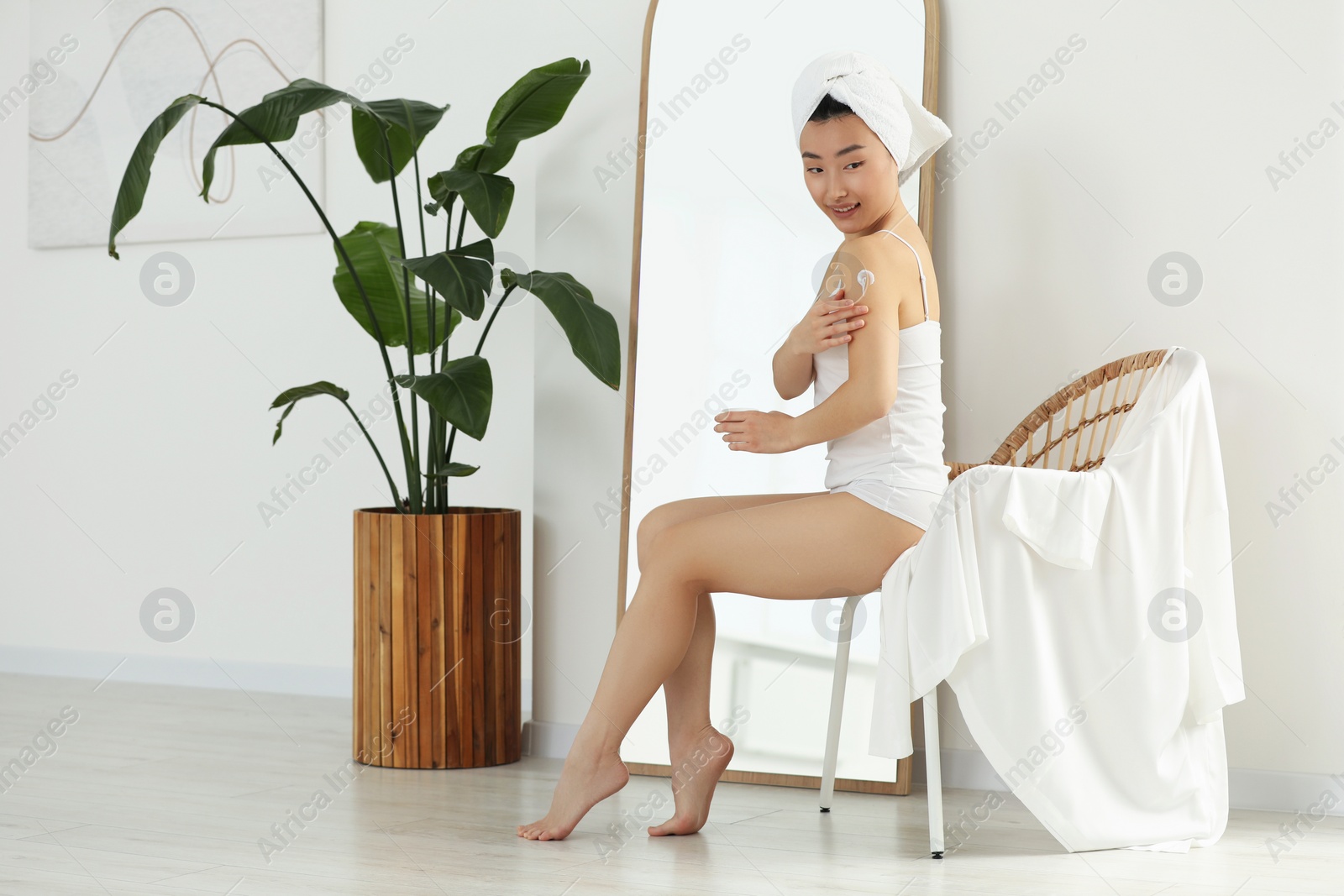 Photo of Beautiful young Asian woman applying body cream on shoulder in room, space for text