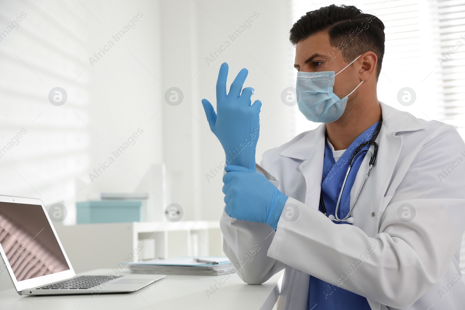 Photo of Doctor in protective mask putting on medical gloves at table in office