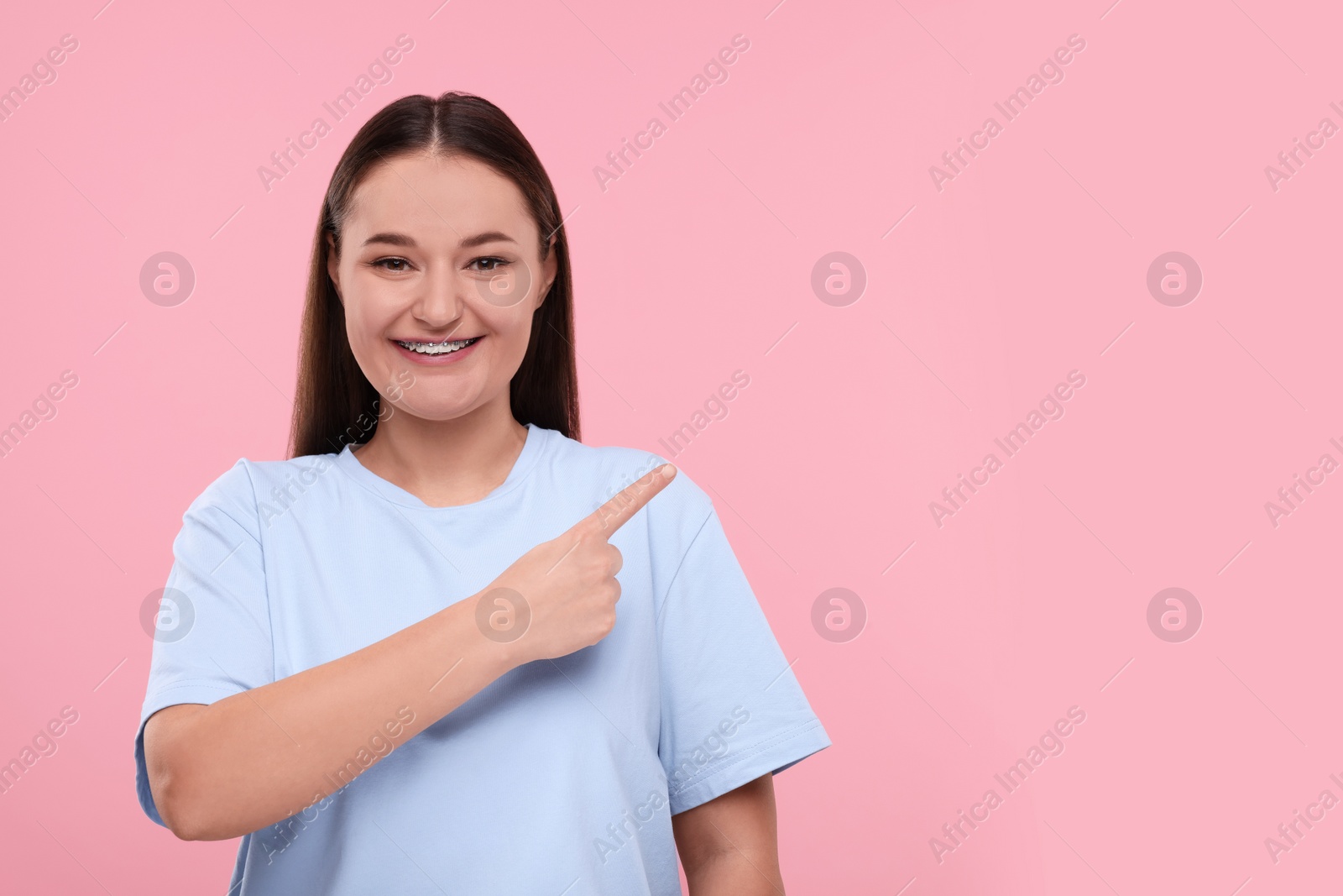 Photo of Smiling woman with dental braces pointing at something on pink background. Space for text