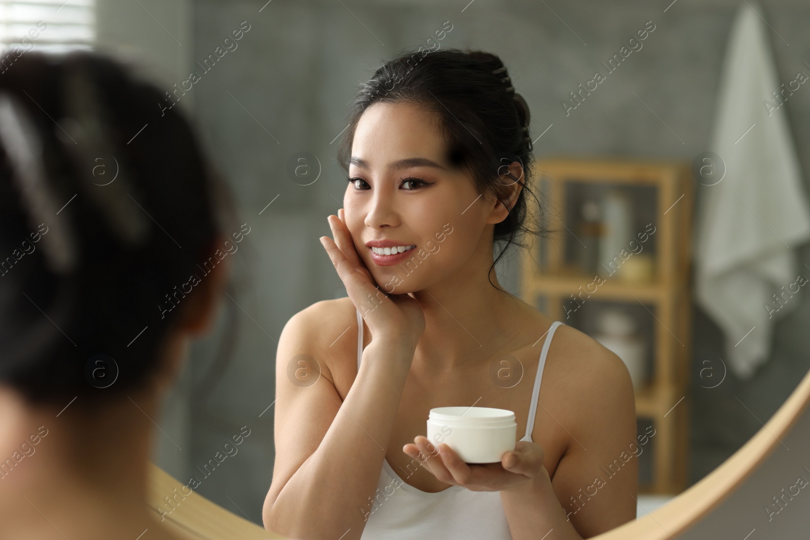 Photo of Happy woman applying face cream near mirror at home