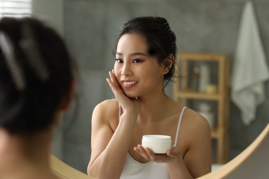 Happy woman applying face cream near mirror at home