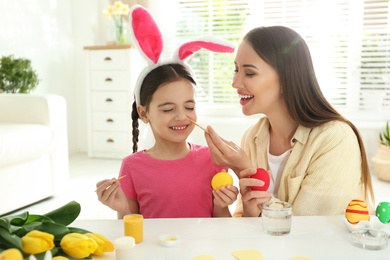 Happy daughter with bunny ears headband and her mother having fun while painting Easter eggs at home