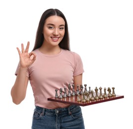 Photo of Happy woman holding chessboard with game pieces and showing OK gesture on white background
