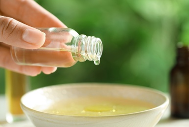 Photo of Woman pouring essential oil from glass bottle into bowl against blurred green background, closeup