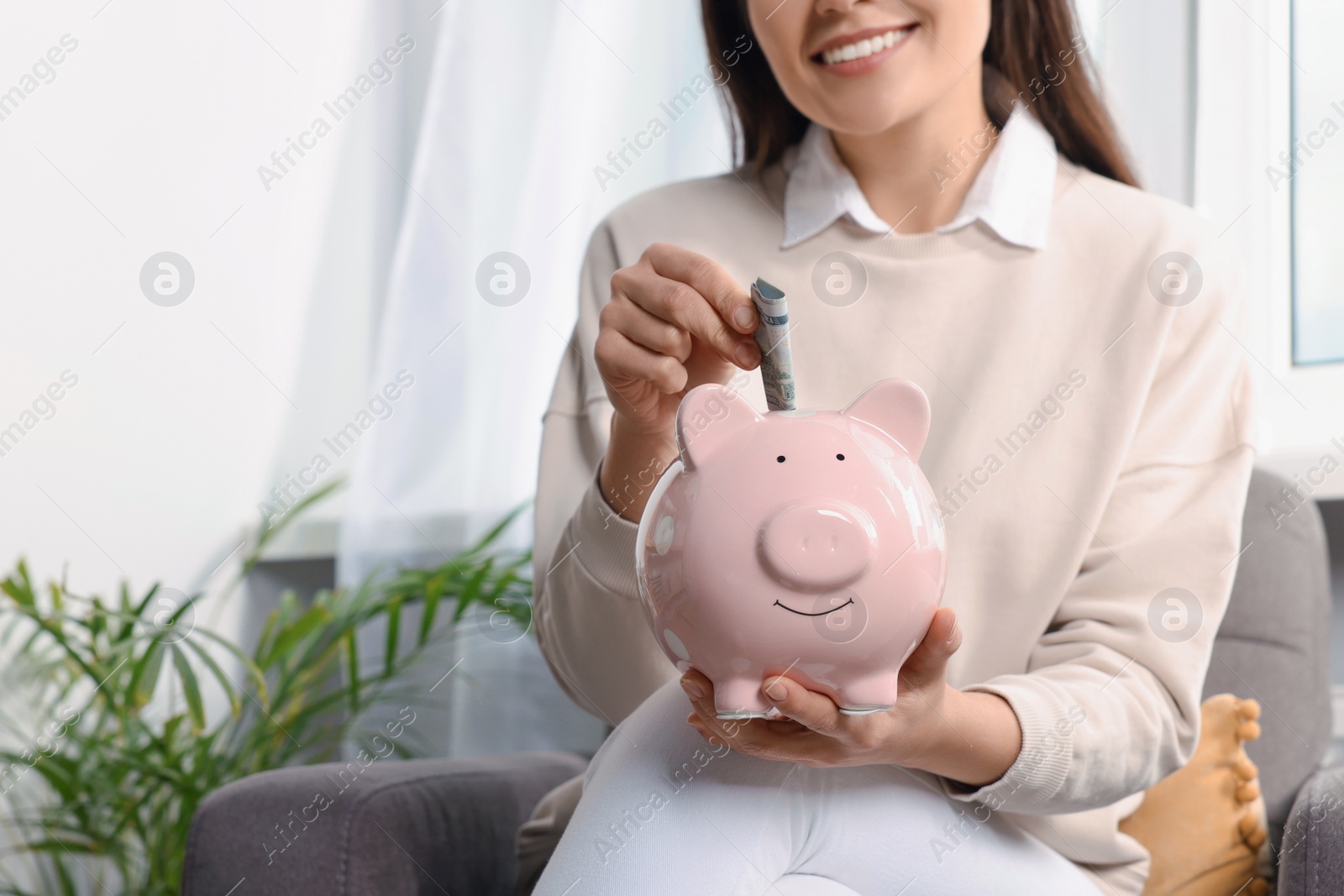 Photo of Happy young woman putting money into piggy bank at home closeup