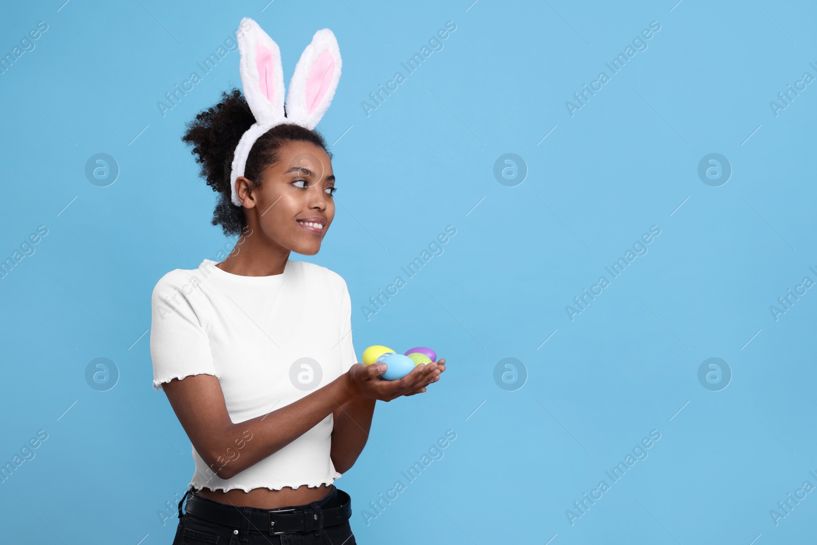 Photo of Happy African American woman in bunny ears headband holding Easter eggs on light blue background. Space for text