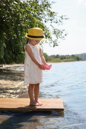 Cute little girl playing with paper boat on wooden pier near river