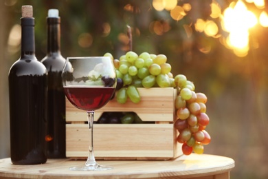 Photo of Bottles and glass of red wine with fresh grapes on wooden table in vineyard