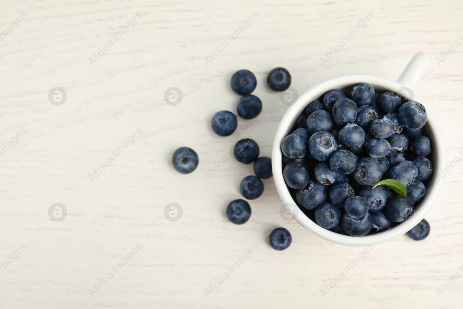 Photo of Fresh ripe blueberries in cup on white wooden table, flat lay. Space for text