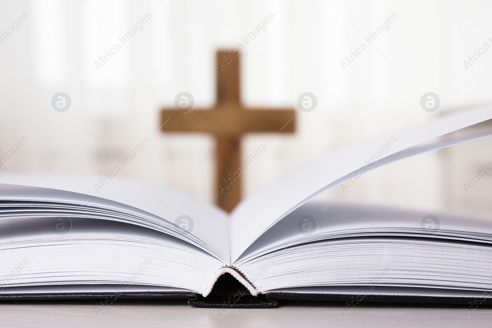 Photo of Open Bible on table and blurred cross on background, closeup. Christian religion