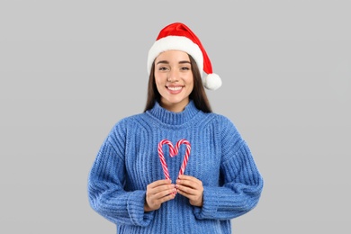 Photo of Young woman in blue sweater and Santa hat holding candy canes on grey background. Celebrating Christmas