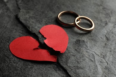 Photo of Halves of torn red paper heart and wedding rings on dark grey table. Broken heart