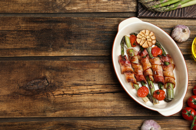 Oven baked asparagus wrapped with bacon in ceramic dish on wooden table, flat lay. Space for text