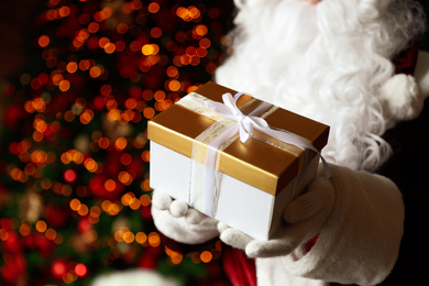 Photo of Santa Claus holding Christmas gift against blurred festive lights, closeup