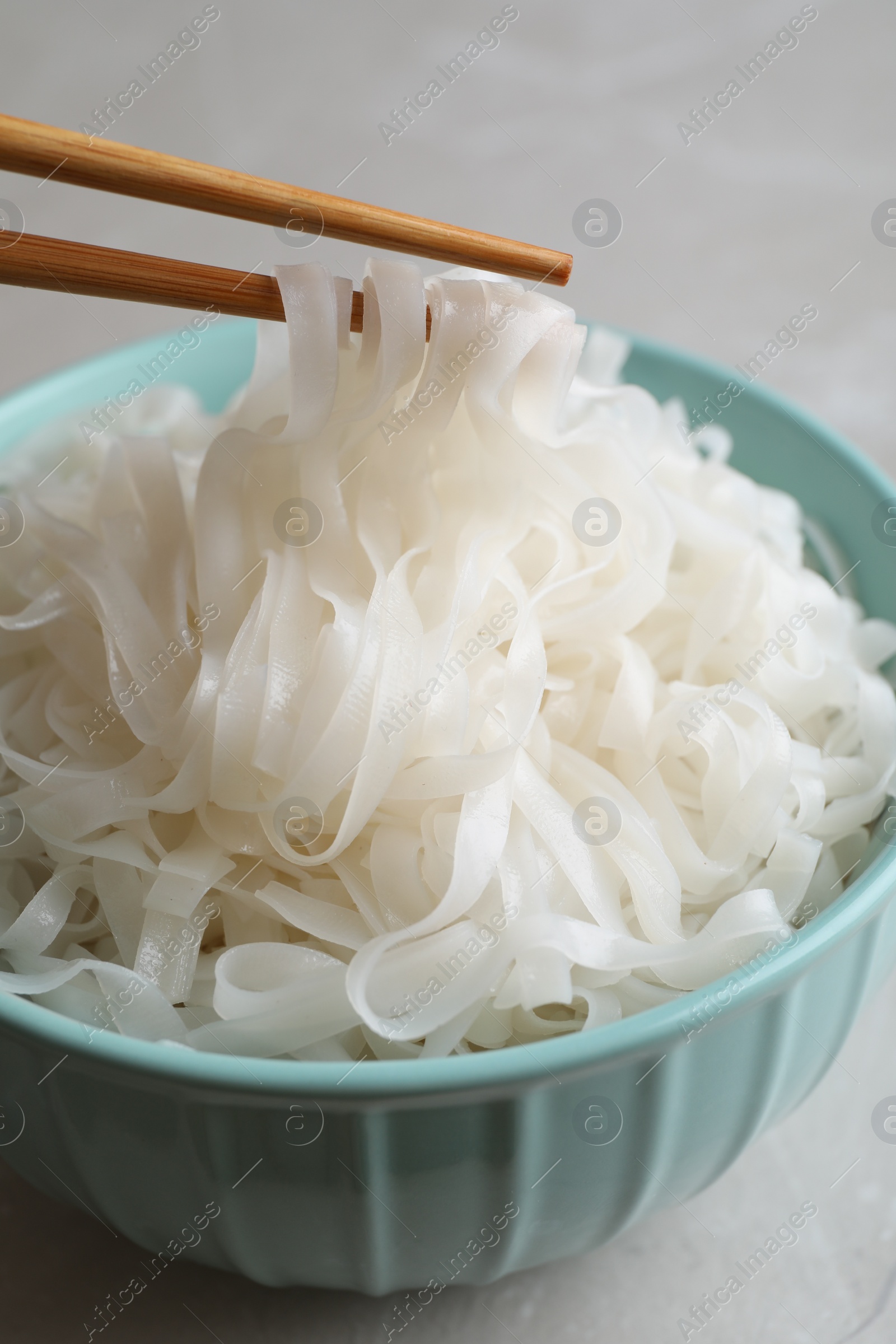 Photo of Taking rice noodles with chopsticks from bowl at light grey table, closeup