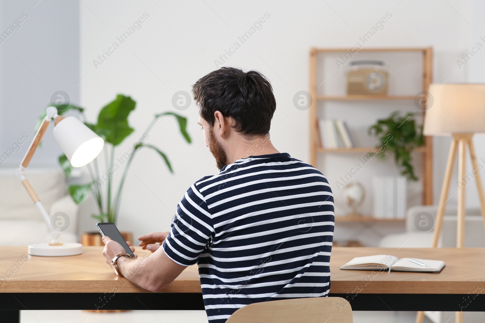 Photo of Home workplace. Man working with smartphone at wooden desk in room, back view