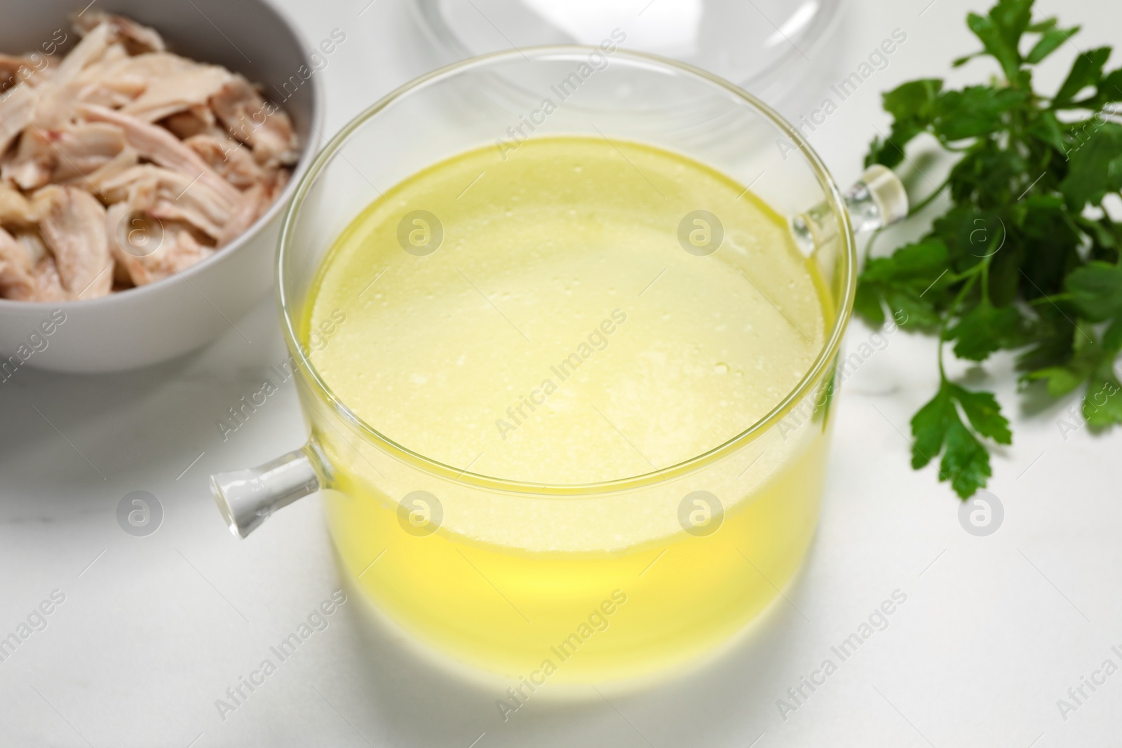 Photo of Glass pot with tasty bouillon, chicken and parsley on white marble table