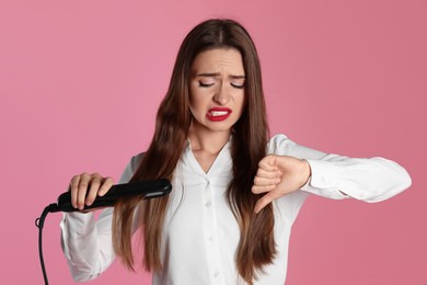Photo of Upset young woman with flattening iron showing thumb down on light pink background. Hair damage