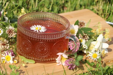 Photo of Ornate glass cup of tea, different wildflowers and herbs on wooden board in meadow, closeup