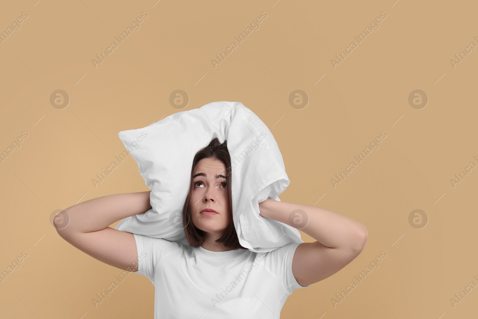 Photo of Unhappy young woman covering ears with pillow on beige background. Insomnia problem