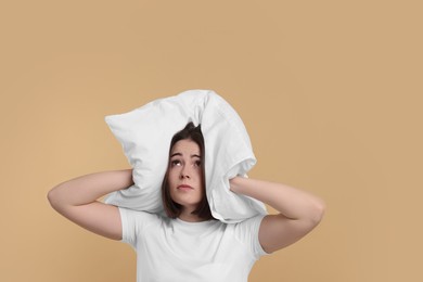 Photo of Unhappy young woman covering ears with pillow on beige background. Insomnia problem