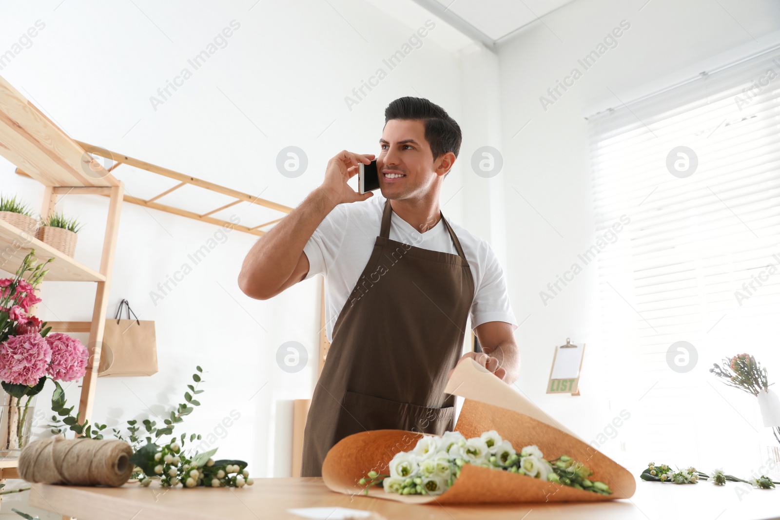 Photo of Florist with beautiful flowers talking on smartphone in workshop