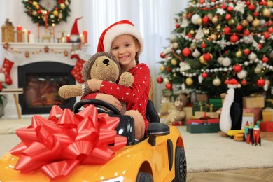 Cute little girl with toy driving children's car in room decorated for Christmas
