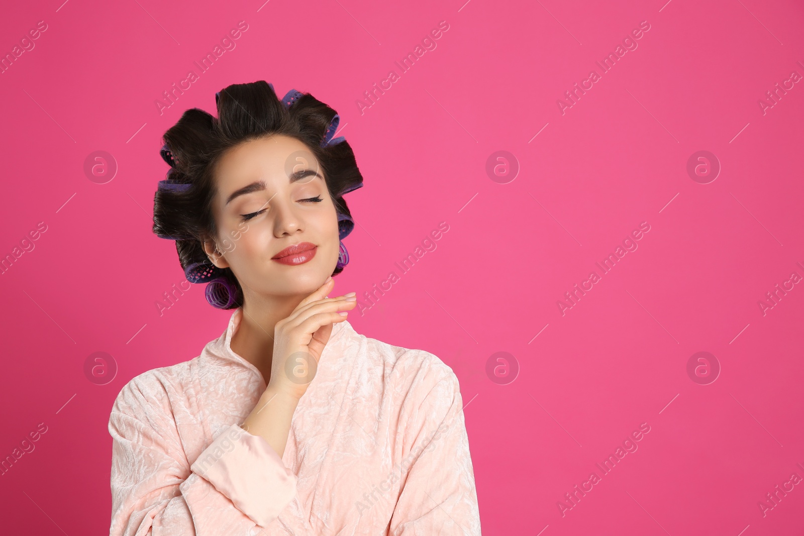 Photo of Happy young woman in bathrobe with hair curlers on pink background, space for text