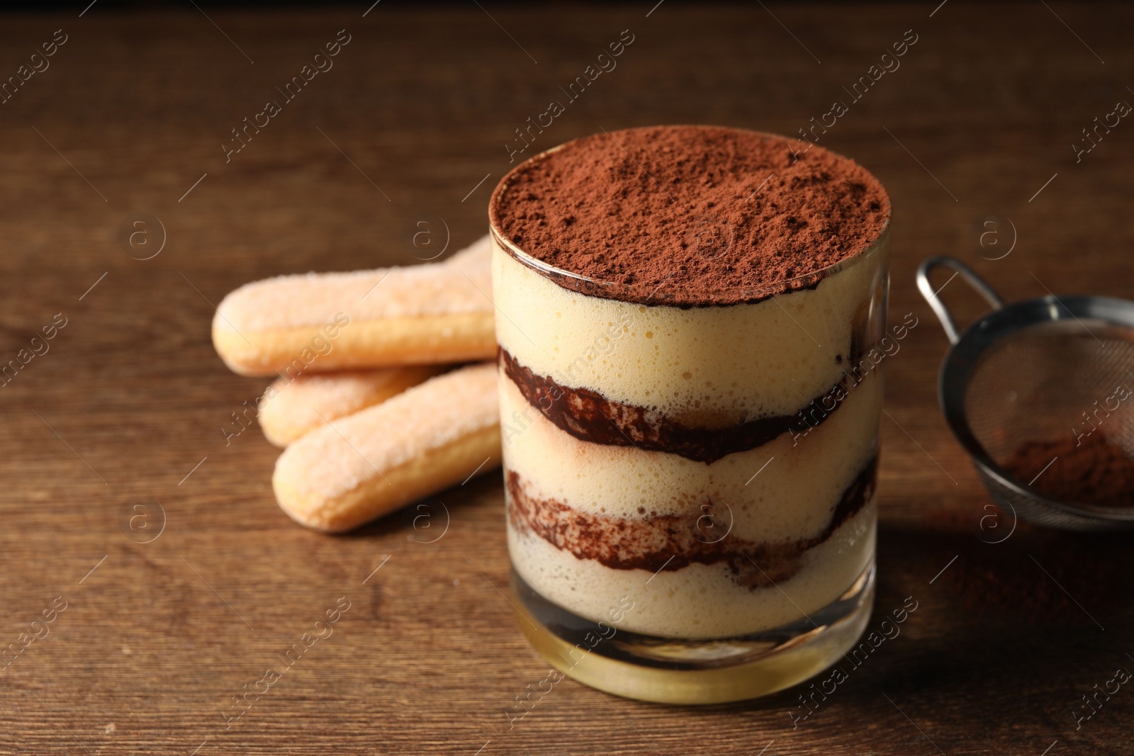 Photo of Delicious tiramisu in glass, biscuits and sieve on wooden table, closeup. Space for text