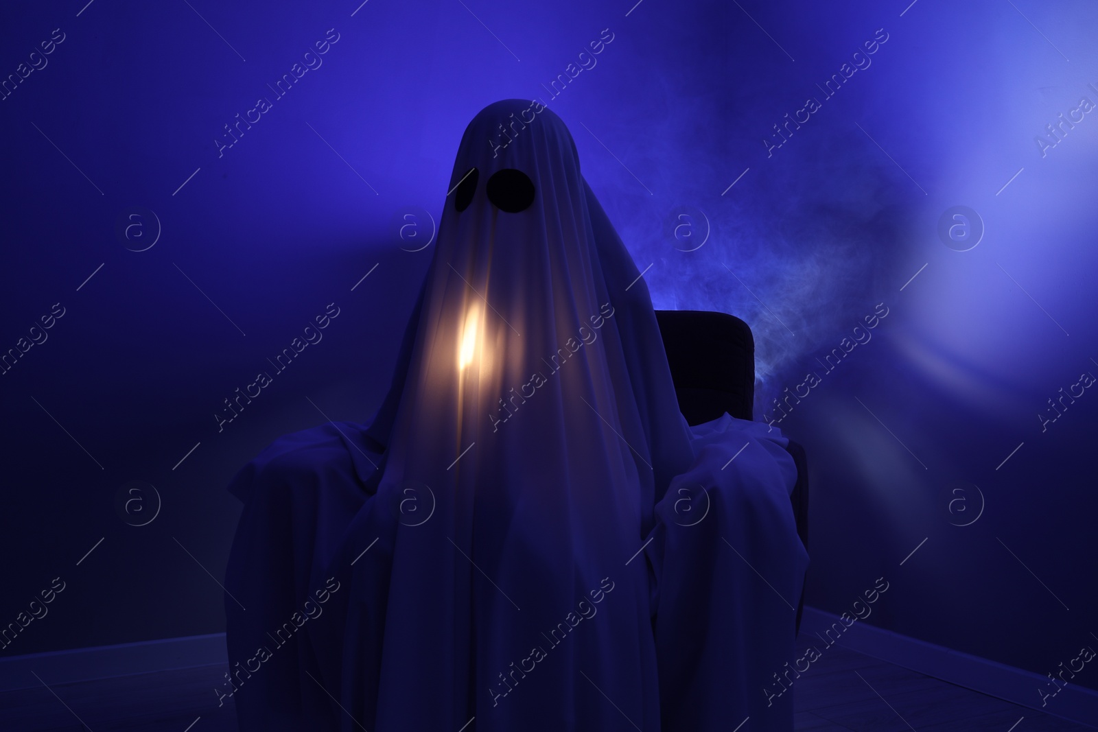 Photo of Creepy ghost. Woman covered with sheet sitting in armchair in blue light