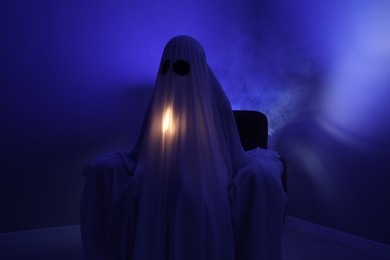 Photo of Creepy ghost. Woman covered with sheet sitting in armchair in blue light