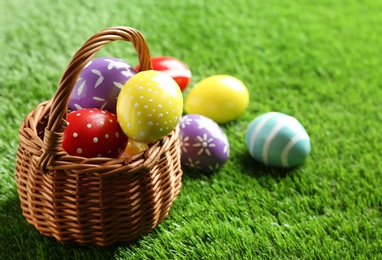 Wicker basket with painted Easter eggs on green grass, space for text