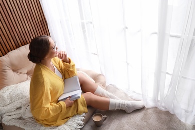 Photo of Beautiful young woman with book and cup of coffee near window at home, space for text. Winter atmosphere