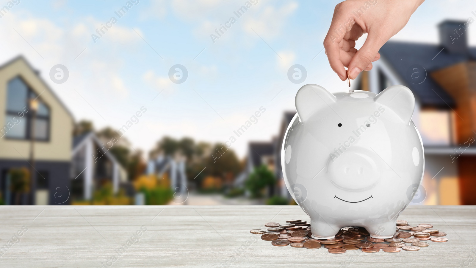 Image of Woman putting coin into piggy bank at white wooden table and blurred view of beautiful houses, space for text. Mortgage concept