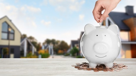 Image of Woman putting coin into piggy bank at white wooden table and blurred view of beautiful houses, space for text. Mortgage concept