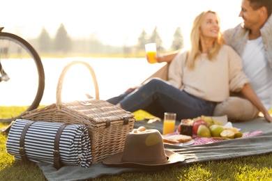 Photo of Happy young couple having picnic near lake, focus on wicker basket with blanket and hat