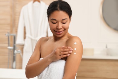 Photo of Young woman applying body cream onto arm in bathroom