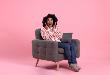Emotional young woman with laptop sitting in armchair against pink background