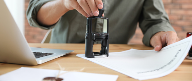 Male notary stamping document at table in office, closeup. Banner design