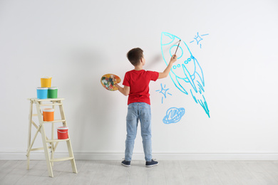 Little child drawing space rocket on white wall indoors