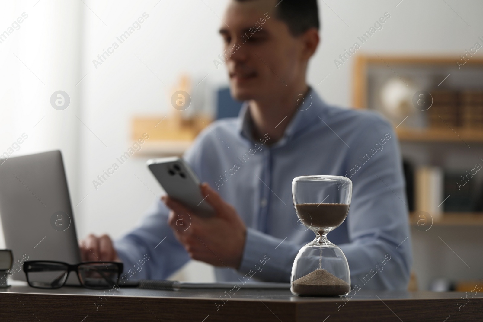 Photo of Hourglass with flowing sand on desk. Man using smartphone while working on laptop indoors, selective focus