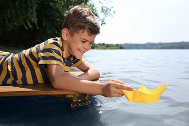 Photo of Cute little boy playing with paper boat on wooden pier near river