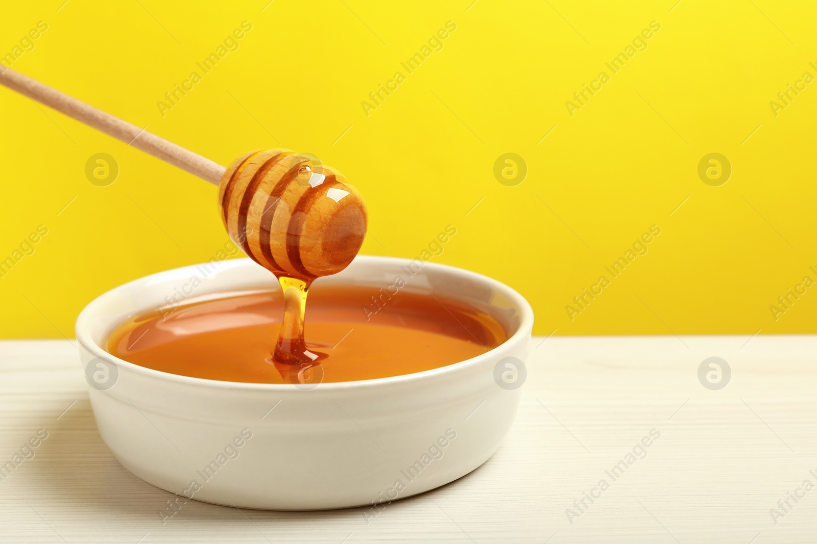 Photo of Pouring tasty honey from dipper into bowl at white wooden table against yellow background, closeup. Space for text