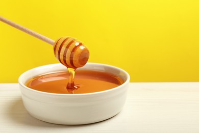 Pouring tasty honey from dipper into bowl at white wooden table against yellow background, closeup. Space for text